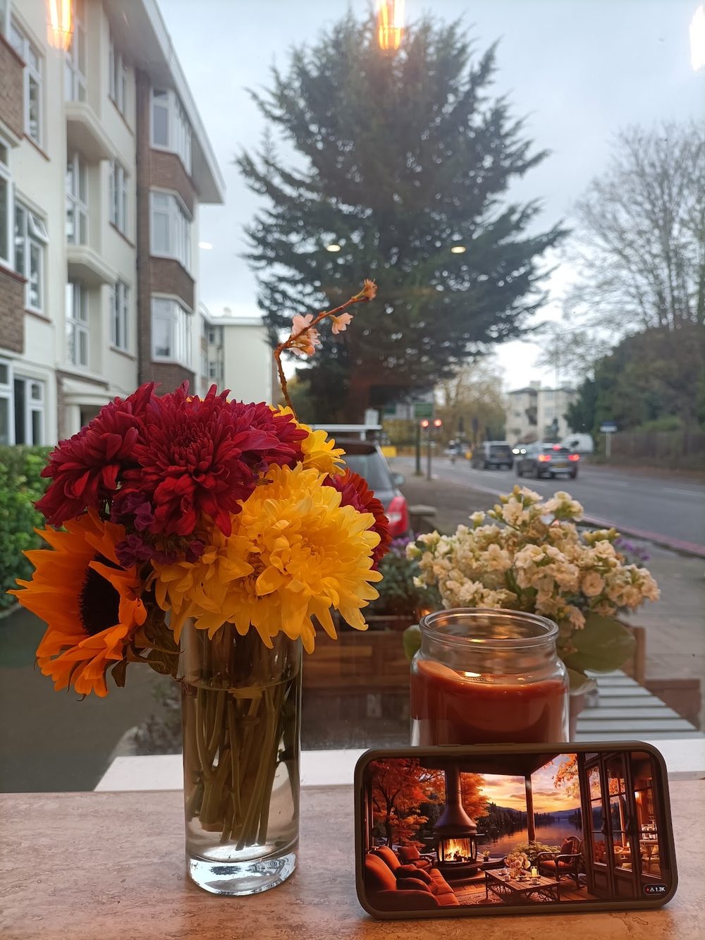 A window-side table with a vase of red, orange and yellow dahlias and a jar candle, looking out onto the street at dusk
